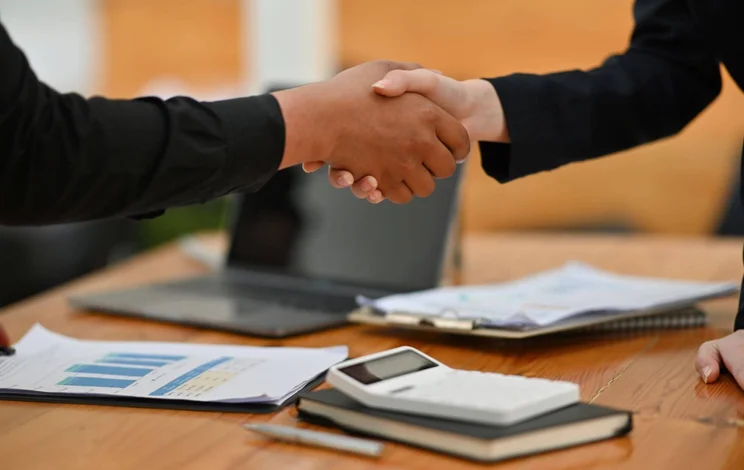 Two people shaking hands over a desk littered with notes and a calculator
