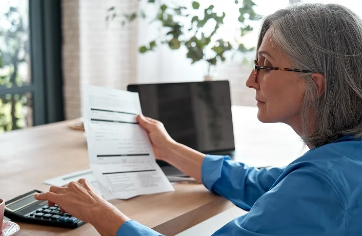 Senior mature business woman holding paper bill using calculator, old lady managing account finances, calculating money budget tax, planning banking loan debt pension payment sit at home office table.
