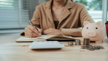 A person writes in a notebook, in front of a calculator and a piggy bank