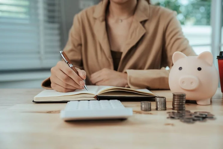 A person writes in a notebook, in front of a calculator and a piggy bank