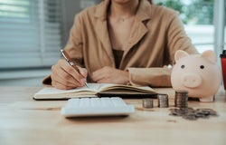 A person writes in a notebook, in front of a calculator and a piggy bank