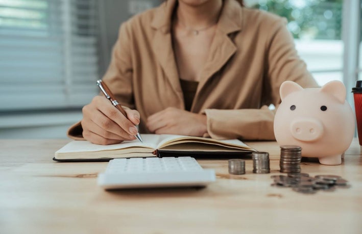 A person writes in a notebook, in front of a calculator and a piggy bank