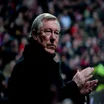 Sir Alex Ferguson watching a football game from the side of the pitch while a crowd of fans watch in the background