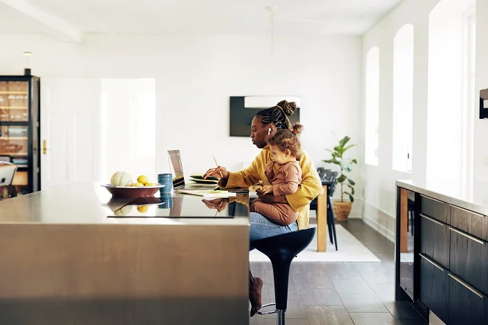 Young African mother working online from home on a laptop with her little daughter sitting on her lap