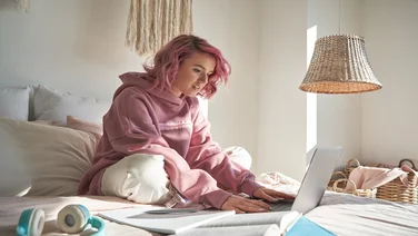 Gen Z girl work employee with pink hair wearing hoodie using laptop computer sitting in bed