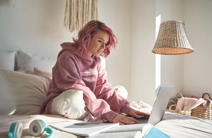 Gen Z girl work employee with pink hair wearing hoodie using laptop computer sitting in bed