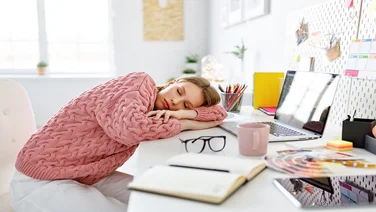 Exhausted young woman lying on table and sleeping while working on remote project in cozy home office