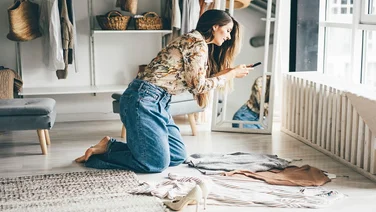 Brunette woman holds smartphone and makes photos of clothes to sell after childbirth via internet. Young woman consults with a designer stylist at home about her wardrobe.