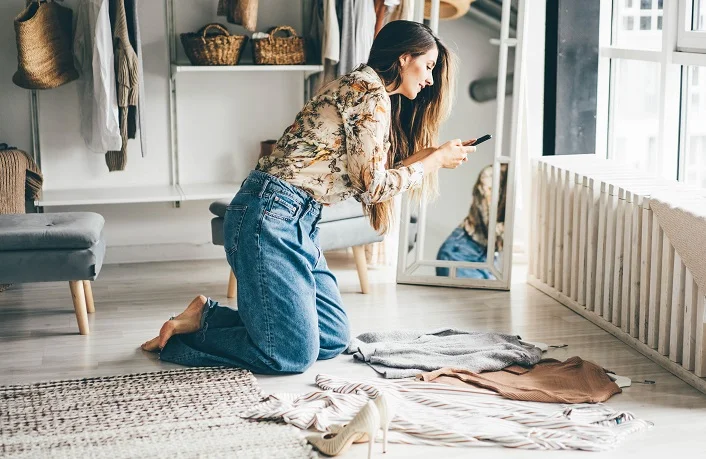 Brunette woman holds smartphone and makes photos of clothes to sell after childbirth via internet. Young woman consults with a designer stylist at home about her wardrobe.