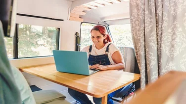 Caucasian young woman working on her laptop in her handmade camper van while traveling. Van road trip holiday and outdoor summer adventure. Nomad remote work lifestyle concept