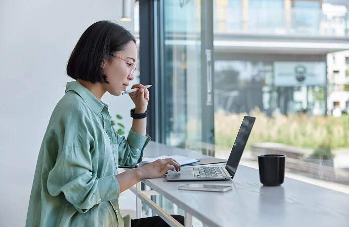 Millennial Asian student prepares for test or exam browses laptop studies in coffee house makes notes in notepad watches online webinar learns foreign language. E learning and online studying concept