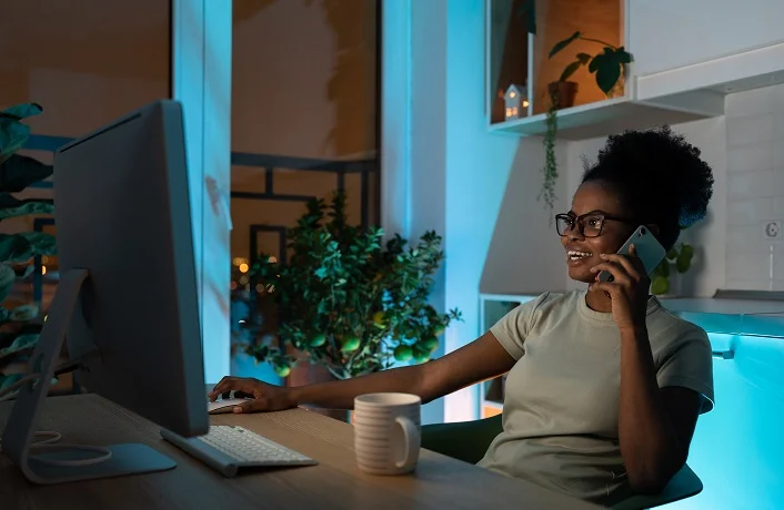 Positive woman in glasses using computer mouse, talking on smartphone, working on desktop pc late at night at home office during the night. Remote work concept
