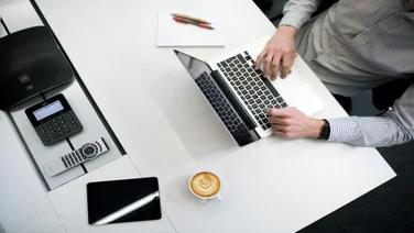 a person typing on their laptop on a white table with coffee and a notepad