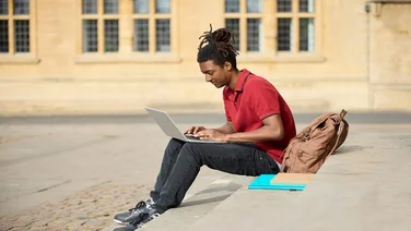 Male Student Working On Laptop Sitting On StepsOutside University Building In Oxford UK