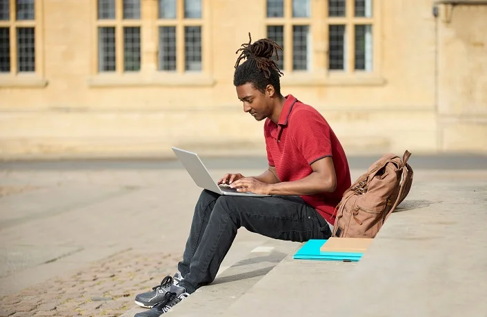 Male Student Working On Laptop Sitting On StepsOutside University Building In Oxford UK