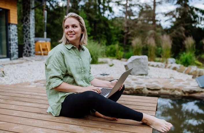 Woman, freelancer working on laptop, sitting on a pier by the backyard lake, a concept of remote office, work during vacation.