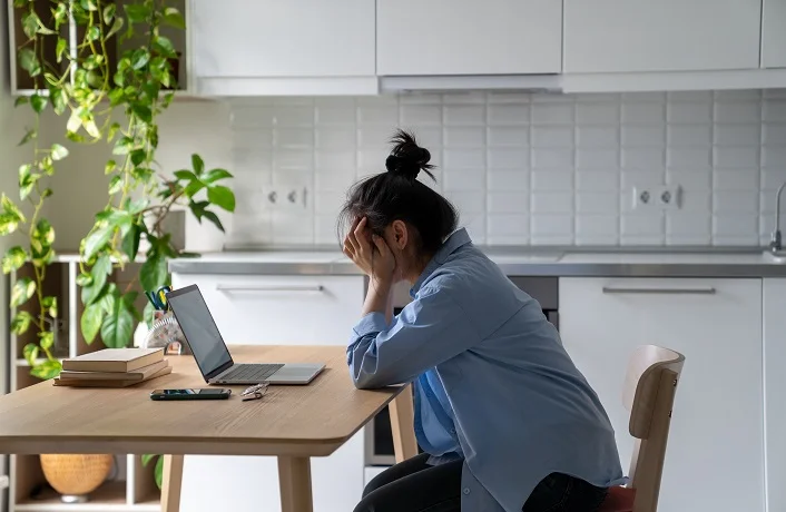 Unhappy young woman sitting at kitchen table looking at laptop having problems in job searching