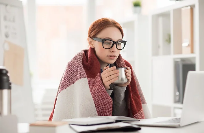 Young woman in plaid and eyeglasses drinking hot tea from mug during work in office