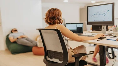 Young, intelligent, business employee working on strategy, innovation, and growth in a dynamic business environment while her male coworker is taking a nap in the bag lying on lazy bag.