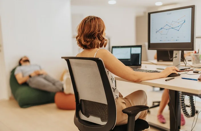 Young, intelligent, business employee working on strategy, innovation, and growth in a dynamic business environment while her male coworker is taking a nap in the bag lying on lazy bag.