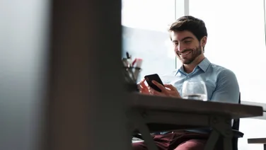 Man sitting at a desk, looking at his smartphone and smiling