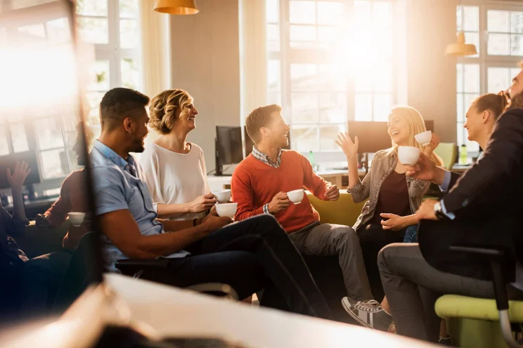 Group of employees sitting around the office chatting and laughing with mugs of coffee.