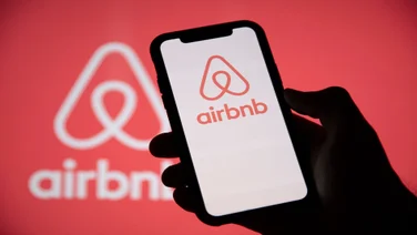 Silhouette of a hand holding up a smartphone displaying the AirBnB logo on its screen against a red backdrop.