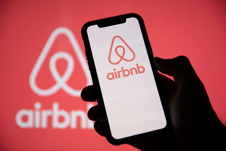 Silhouette of a hand holding up a smartphone displaying the AirBnB logo on its screen against a red backdrop.