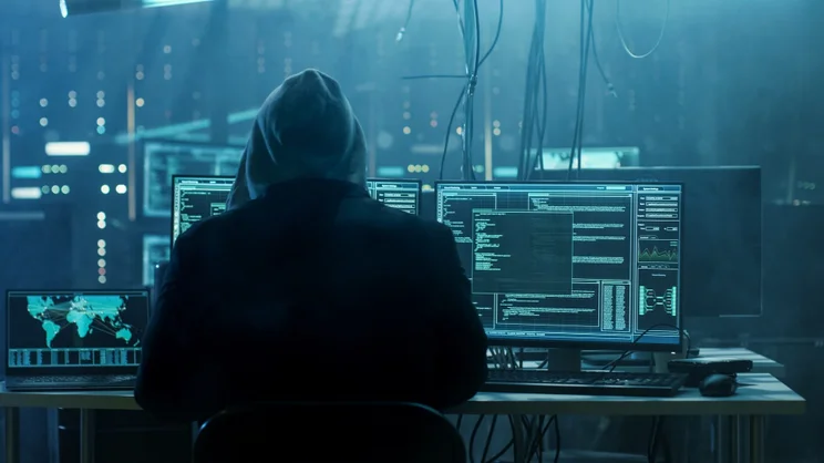 Backshot of a hooded person in a dark room with multiple computer screens.