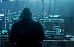 Backshot of a hooded person in a dark room with multiple computer screens.