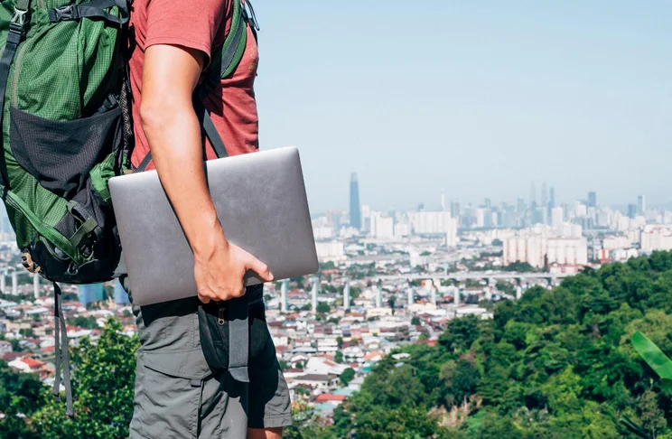 A man wearing a red shirt, cargo shorts and large green backpack walking along the cityscape with a laptop in his right arm.