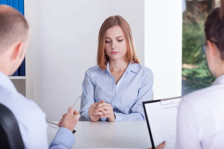 A young woman in a light blue shirt sitting at a table facing two interviewers. She is looking down at her hands and appears uncomfortable.