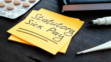 A yellow post it note reading "Statutory Sick Pay" stuck on a black wooden desk.