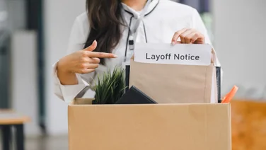 A woman with a white shirt standing behind a box of office supplies holding an envelope that reads "layoff notice"
