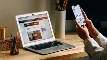 A person uses their smartphone to check something, while their laptop sits open on their desk