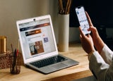 A person uses their smartphone to check something, while their laptop sits open on their desk