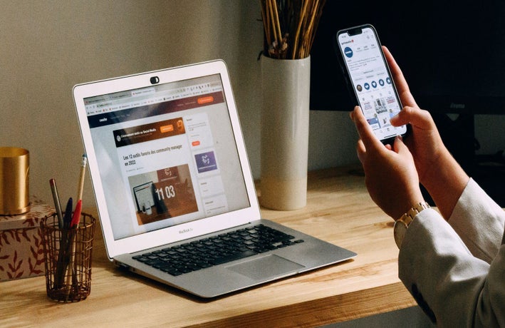 A person uses their smartphone to check something, while their laptop sits open on their desk
