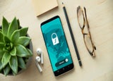 A smartphone, pencil, earbuds, and a pair of glasses are laid out on a desk. The smartphone displays a graphic of a padlock.