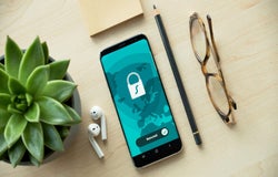 A smartphone, pencil, earbuds, and a pair of glasses are laid out on a desk. The smartphone displays a graphic of a padlock.