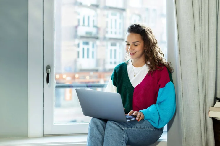 A smiling woman sits by a window working on her laptop.