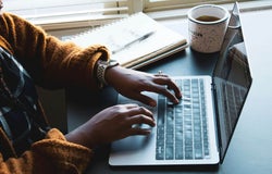 A person sits working at their laptop with a notebook and a hot drink.
