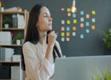A woman sits in front of her laptop, deep in thought, behind a wall of sticky notes.