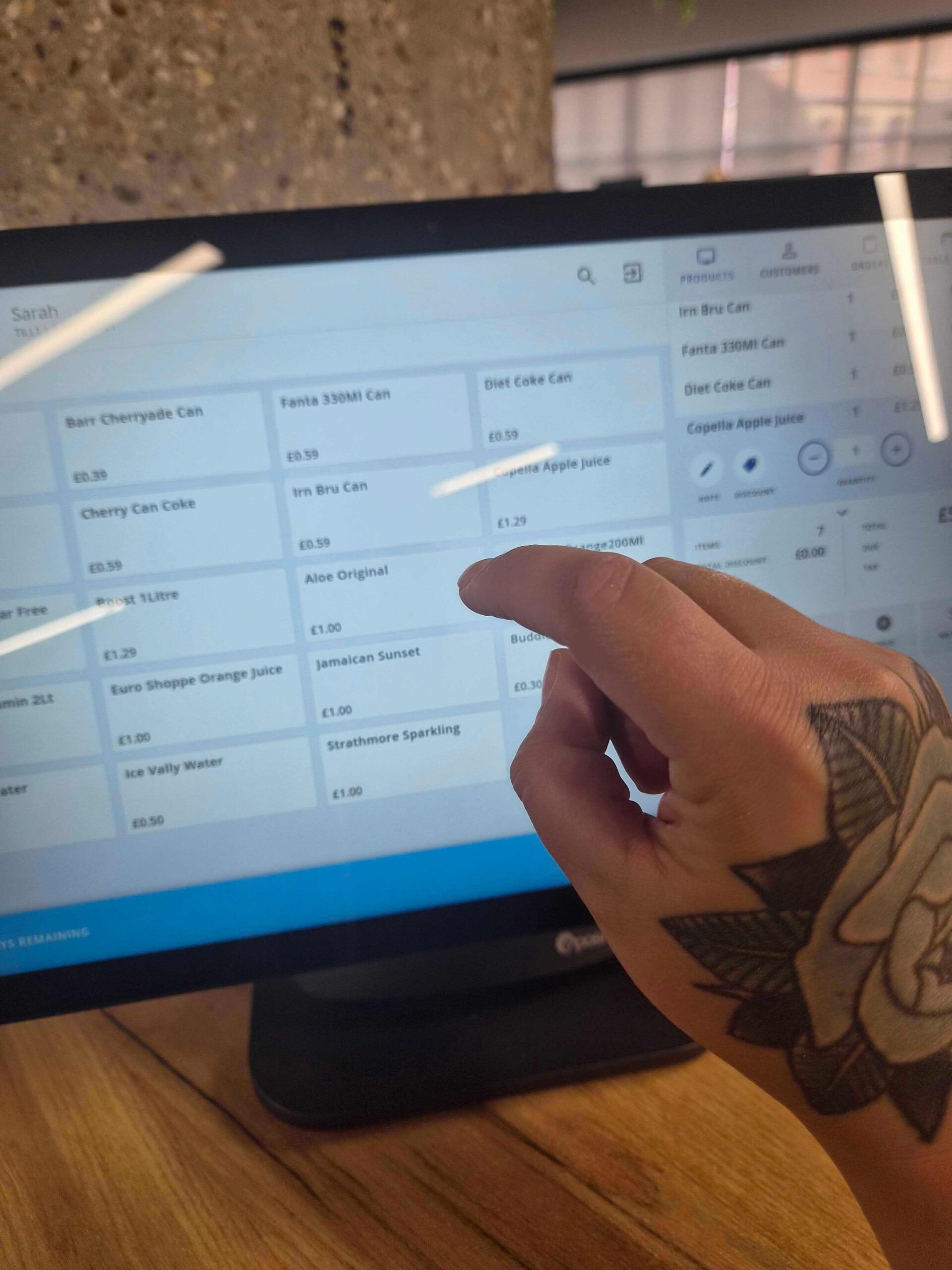 A close-up view of a person with a flower tattoo on their hand reaching out to touch the Epos Now Countertop 2 touchscreen POS screen displaying soft drink product options.
